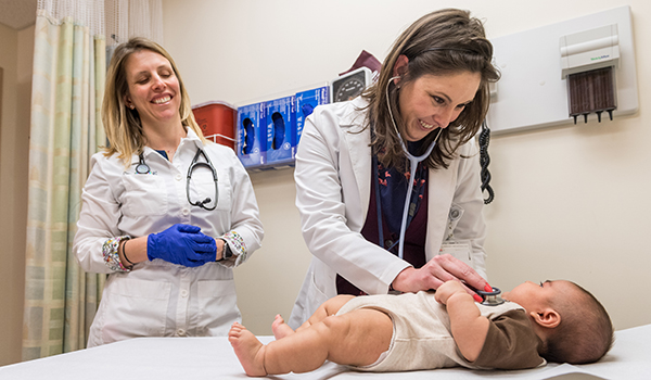 Michigan nurse examines baby with stethoscope. Fellow nurse stands behind her and smiles.